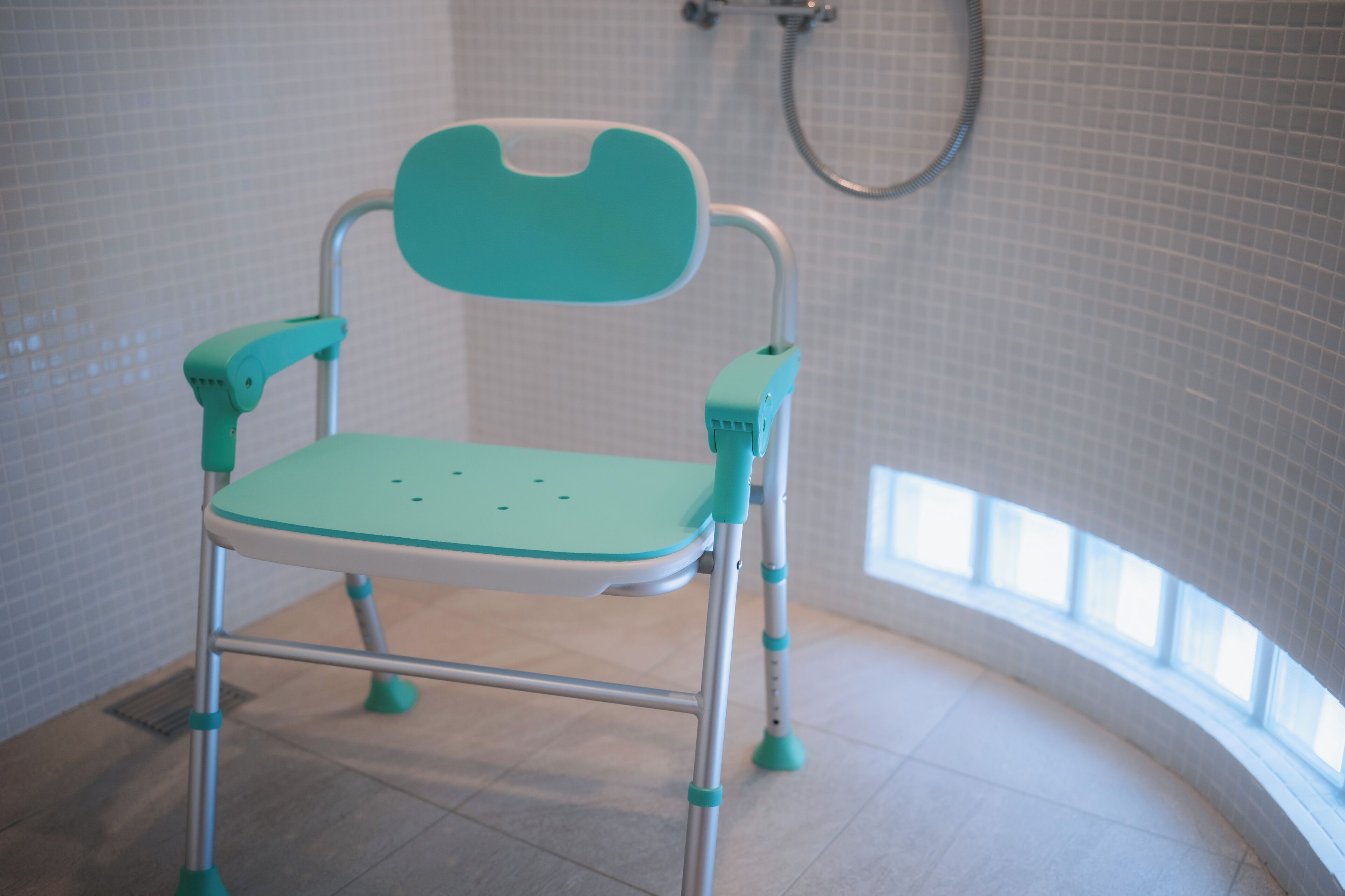 A teal-colored child’s high chair in a bathroom with tiled walls and a shower.