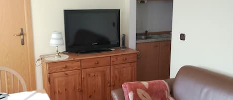 Un salon chaleureux avec un canapé en cuir marron, une table à manger en bois avec une chaise, un téléviseur posé sur un meuble en bois, et un coin cuisine visible à travers une porte ouverte.