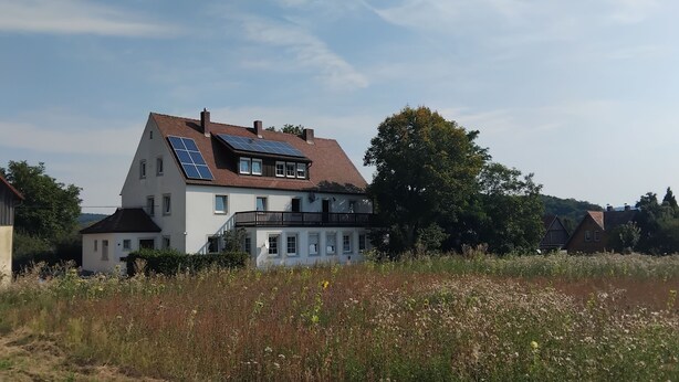 Uma casa de dois andares com painéis solares no telhado, cercada por um campo de flores silvestres e árvores.