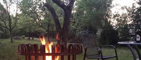 Un hogar de fuego metálico con llamas, rodeado de césped y árboles.