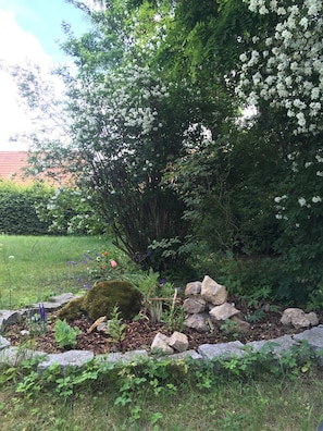 Un jardín con diversas plantas, que incluye un árbol de flores blancas, una roca cubierta de musgo y una pequeña construcción de madera.
