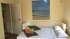 Ett sovrum med en säng, en dörr, en tavla med ett strandmotiv som hänger på väggen och en termostat.