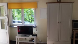 Ett rum med en tv på ett vitt tv-bord, ett fönster med en gul persienn, en vit garderob och ett litet bord med elektroniska apparater.
