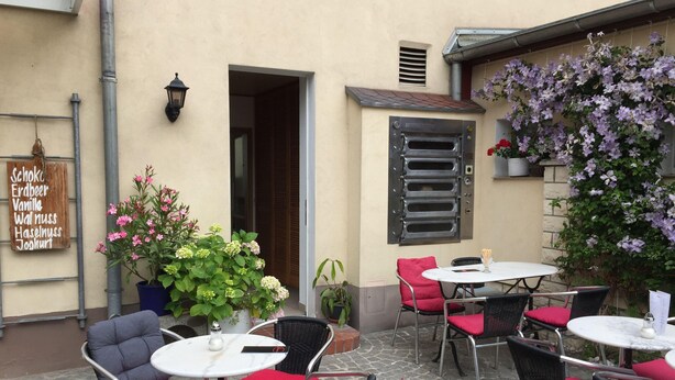 An outdoor cafe area with tables and chairs, a sign listing food items, and potted plants. - Kleine Mietidee (Markranstaedt)