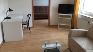 A compact living space with a white desk, a black chair, a brown sofa, a television on a white stand, and a glass coffee table. - Kleine Mietidee (Markranstaedt)