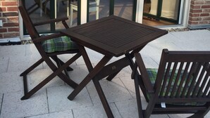 An outdoor patio with a wooden table and chairs, a brick building, and a glass door leading to an interior space. - Huus Windroos Wohnung 2 (Jemgum)