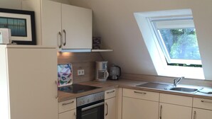 A kitchen with white cabinets, a built-in oven, a coffee maker, and a window with a view of trees. - Huus Windroos Wohnung 2 (Jemgum)