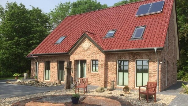 A brick house with a red roof, solar panels, and a circular gravel area with a bench and a potted plant. - Huus Windroos Wohnung 2 (Jemgum)