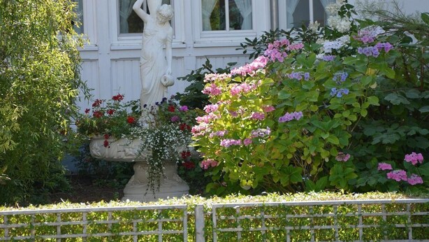 Um edifício branco com uma estátua na frente, cercado por vegetação exuberante e plantas floridas.