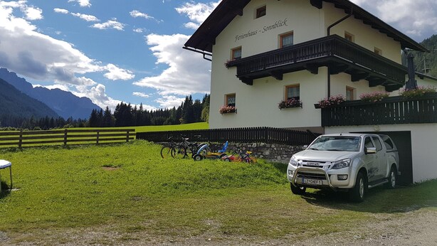 En traditionell byggnad i alpmiljö med en balkong, en parkerad bil och en bergskedja i bakgrunden.