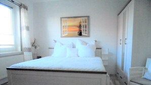 A bedroom with a large bed, a window with curtains, a painting on the wall, and a chair. - Ferienwohnungen Seidel (Zingst)