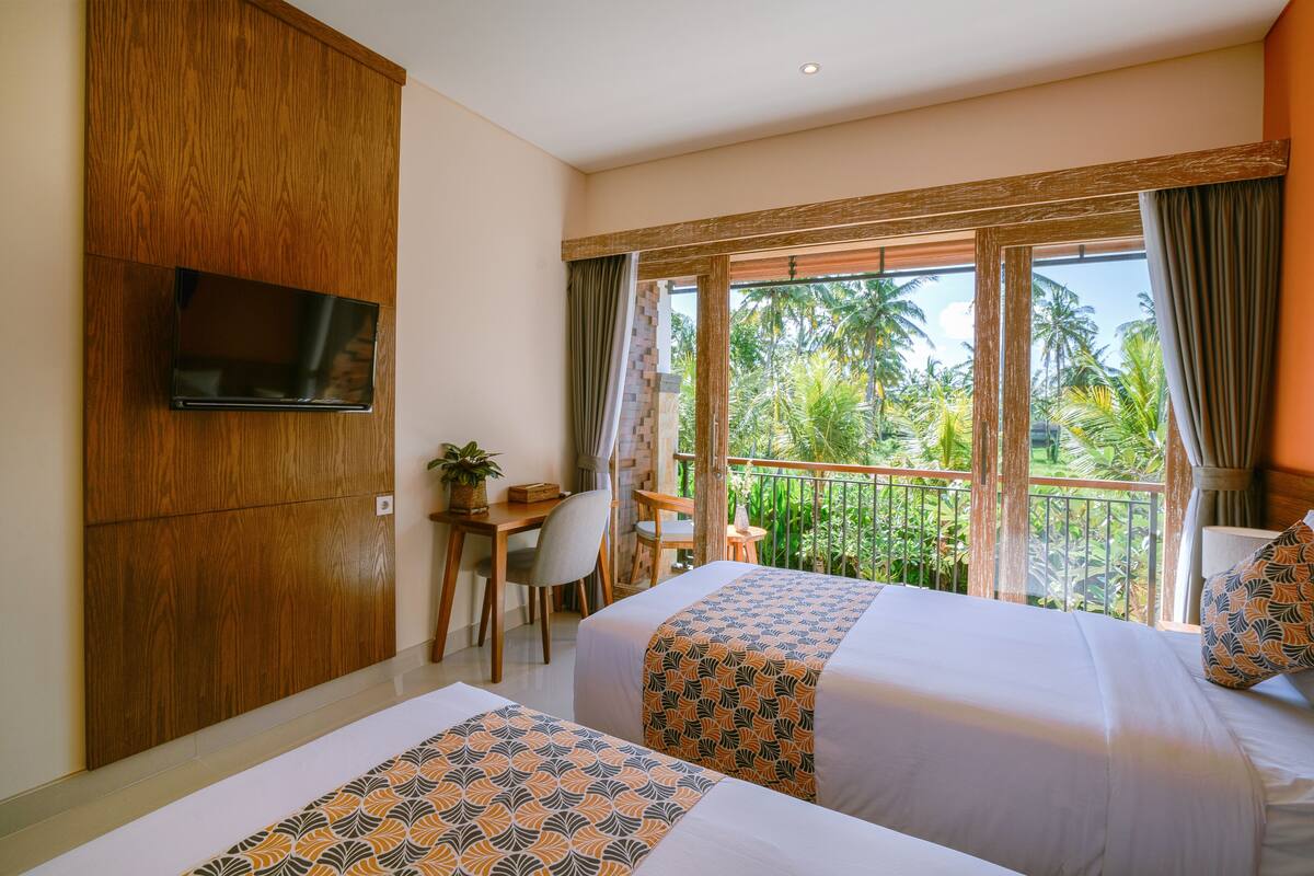 A hotel room with two beds, a desk, a TV, and a balcony with a view of palm trees.