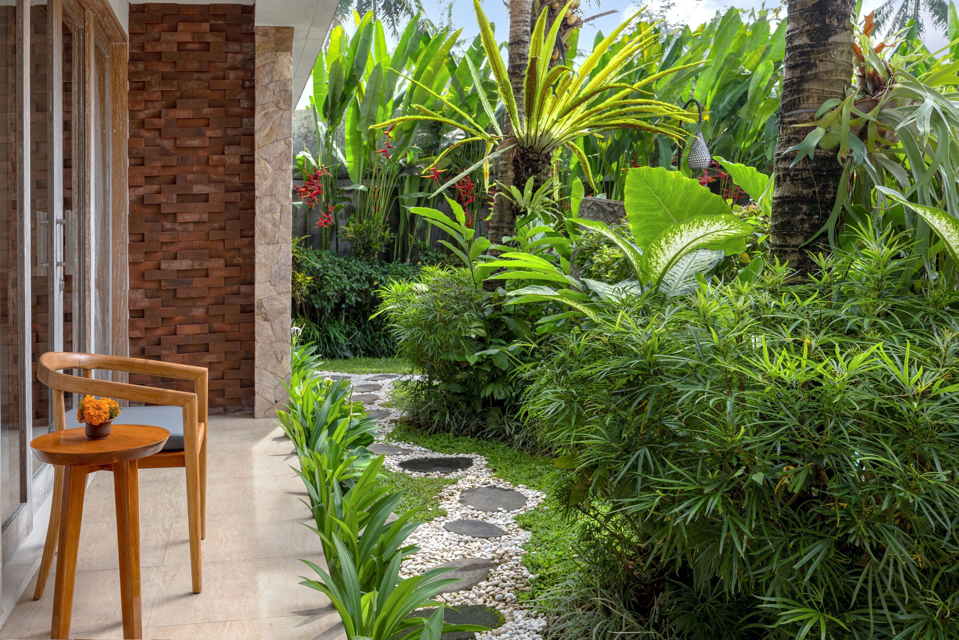 A garden pathway with a wooden chair and table, surrounded by lush greenery and a brick wall.