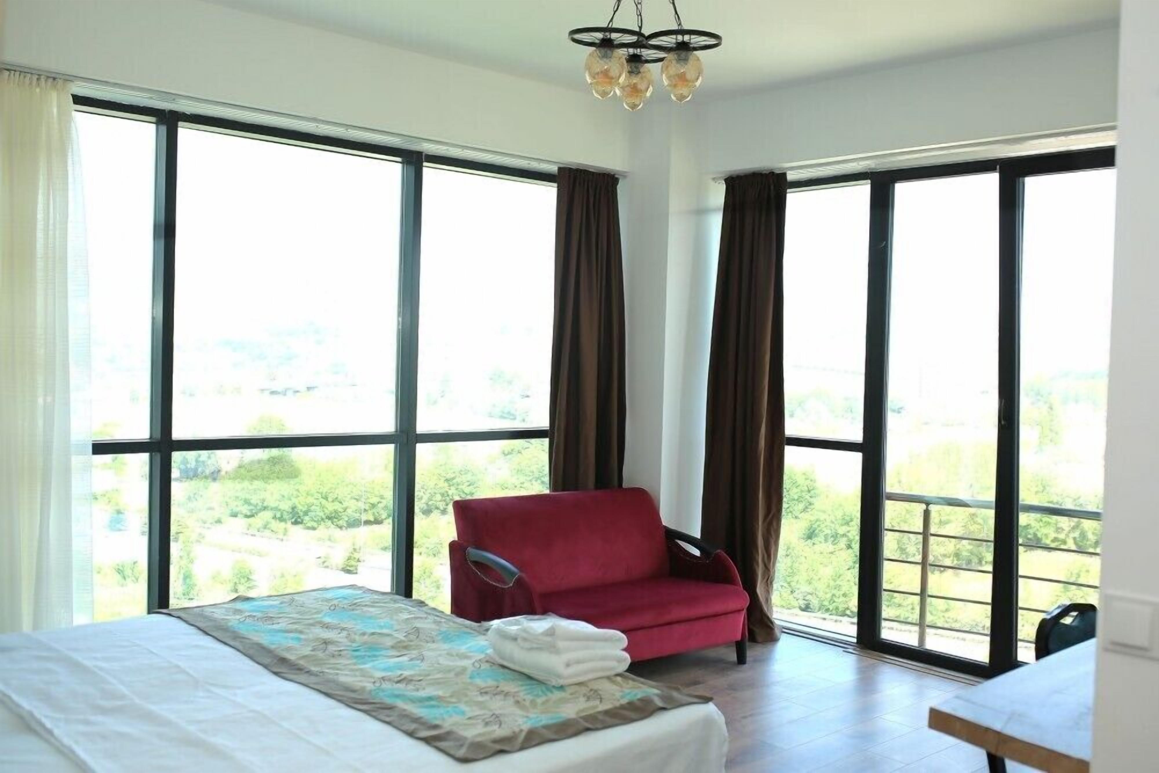 A hotel room with a large bed, a red armchair, and a view of greenery through large windows.