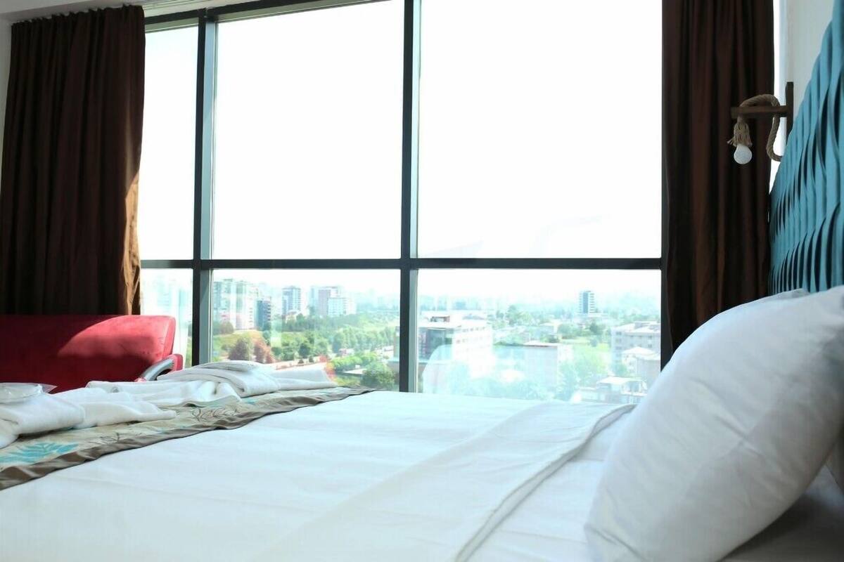 A hotel room with a large window, a bed with white linens, a red chair, and a view of a cityscape.