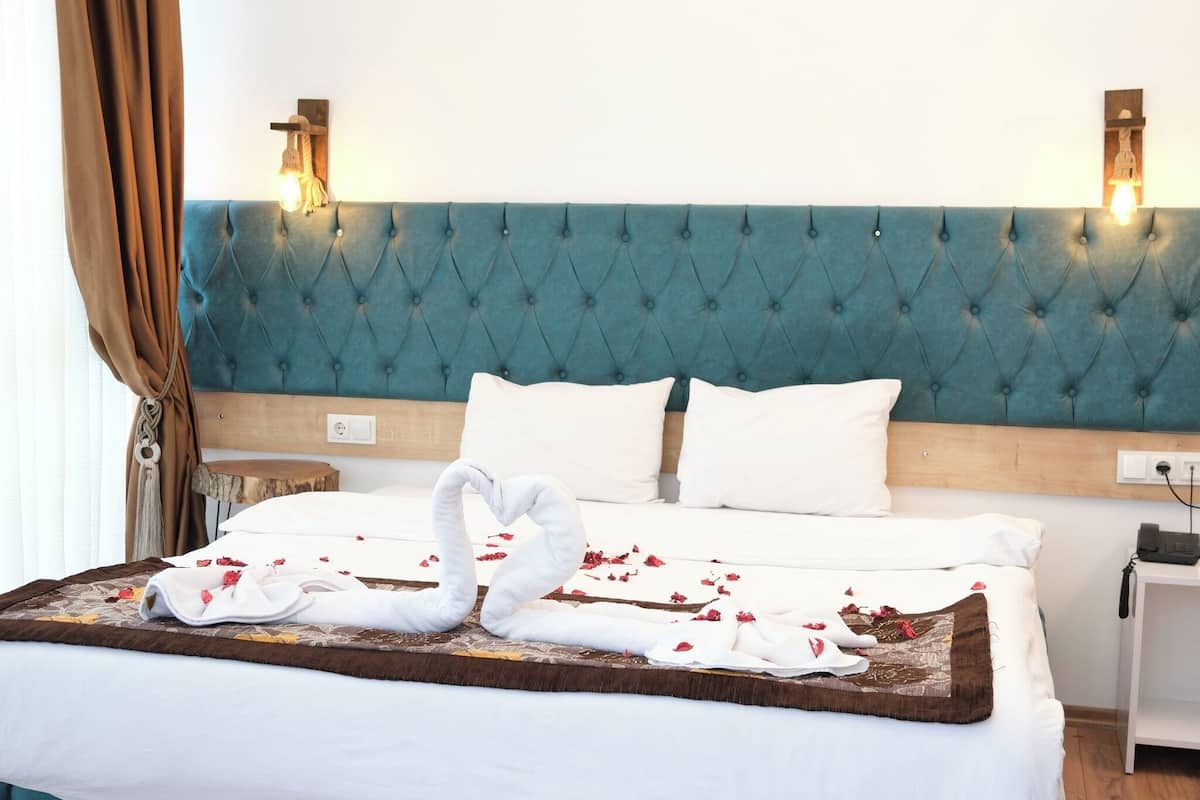 A hotel room with a tufted teal headboard, white bedding, a swan-shaped towel arrangement, and a wooden nightstand with a phone.