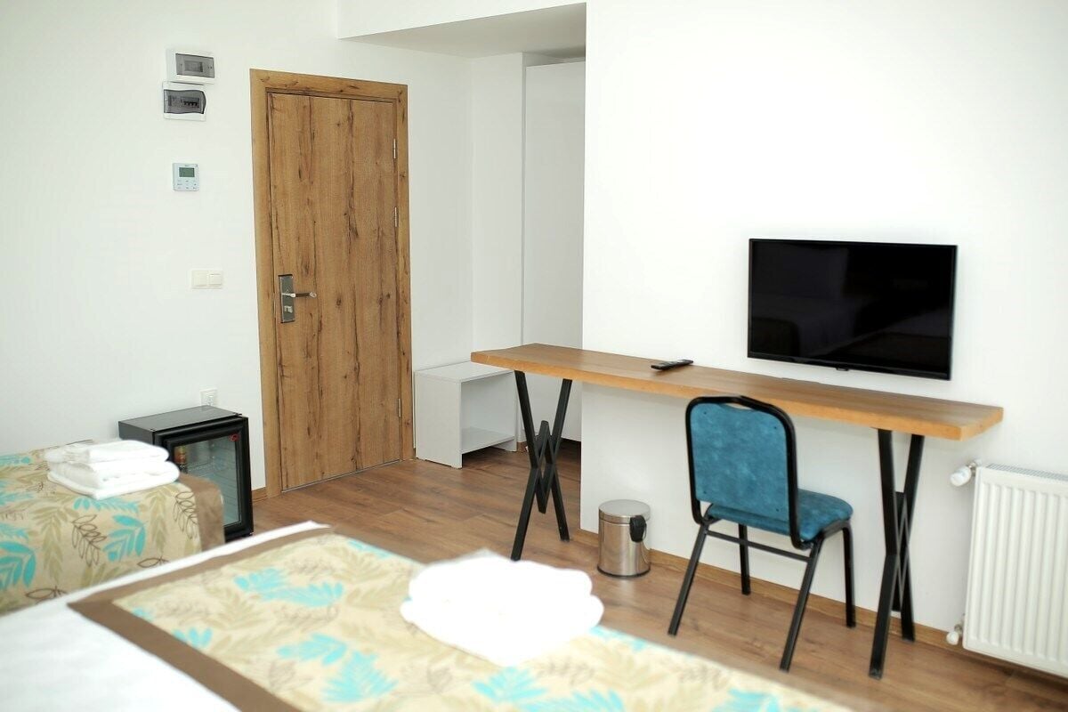 A room with a wooden door, a TV mounted on the wall, a desk with a chair, a bed with a patterned bedspread, and a radiator.