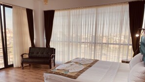 Habitación de hotel con cama, una silla, mesita de noche con teléfono y control remoto, y un ventanal con cortinas.