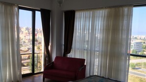Habitación de hotel con una cama grande, un sofá rojo y vistas de un paisaje urbano a través de amplios ventanales.