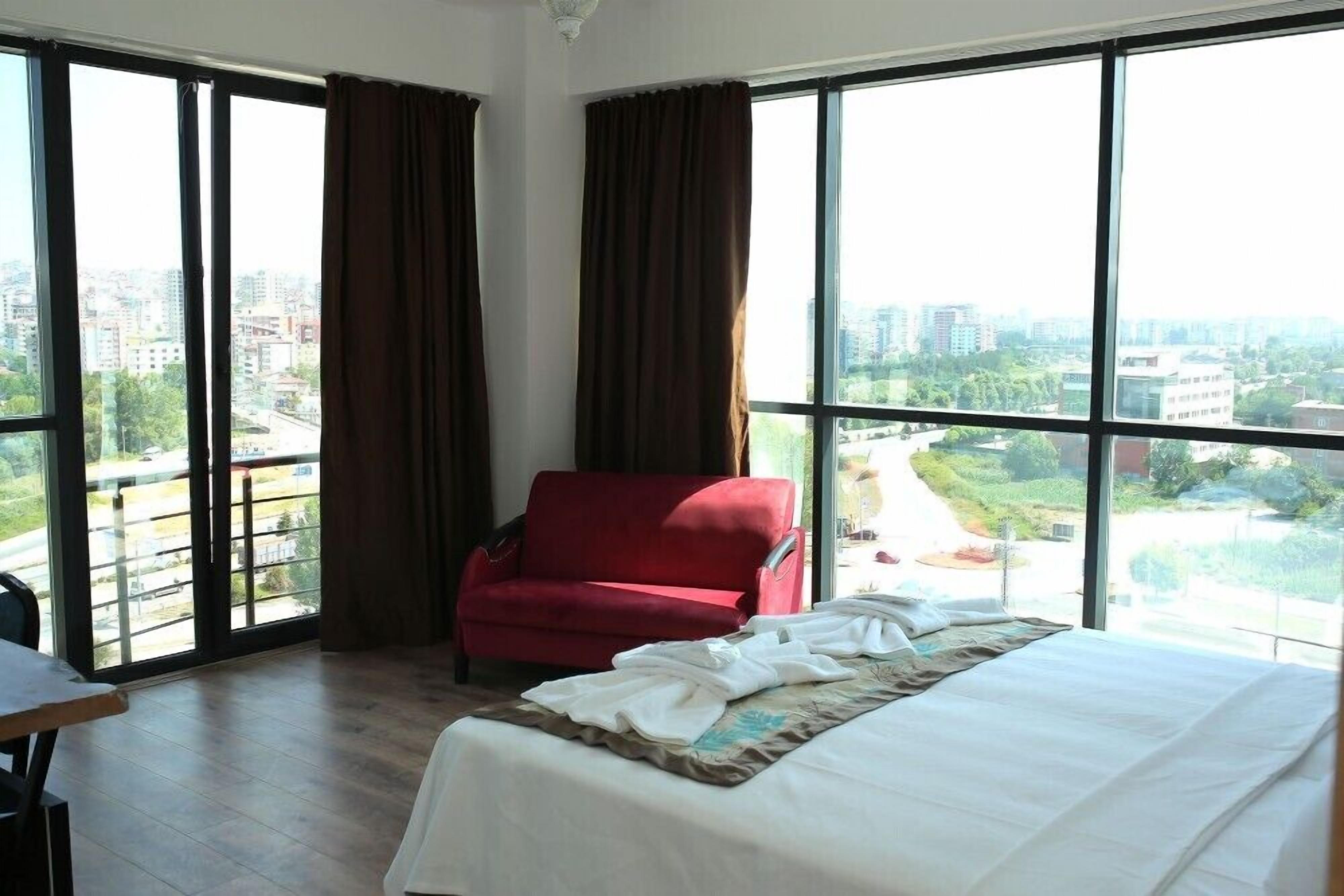 A hotel room with a large bed, a red armchair, and a view of a cityscape through large windows.