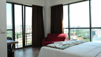A hotel room with a large bed, a red chair, a dining table, and large windows offering a city view.