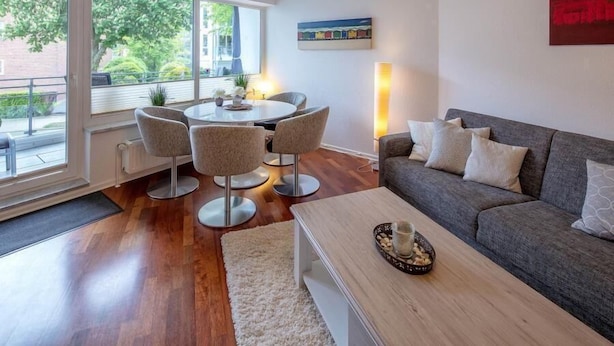 A modern living room with a grey sofa, a round dining table with four chairs, and a large window offering a view of the outdoors. - Residenz Barke App 10 (Scharbeutz)