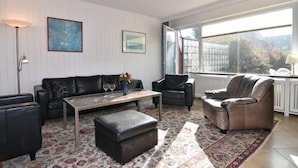 Ein Wohnzimmer mit einer Couch, Sesseln, einem Couchtisch und einem Teppich. Ein Fenster bietet Blick auf Gebäude und Bäume.