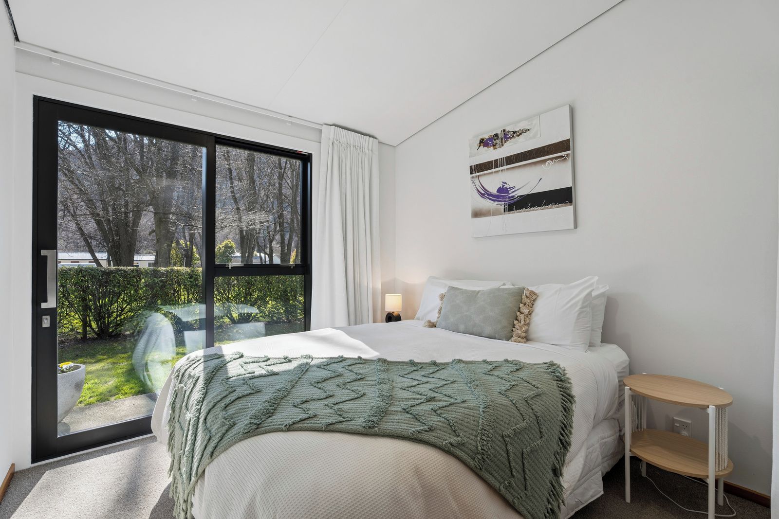 A bedroom with a bed, a bedside table, a nightstand, a window, and a view of the outdoors.