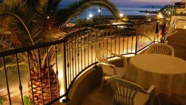Un balcón con vista al mar al atardecer, con una palmera, una mesa con sillas y una barandilla.