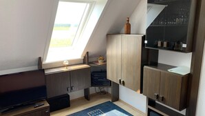 A room with a sloped ceiling, a TV on a wooden stand, a cabinet with a bottle on top, and a shelf with glassware. - Ferienhaus Hirschrudel - Ferienwohnung 3 (Katzow)