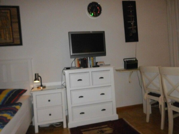 Un dormitorio con cama, un tocador blanco, televisor, reloj y una silla.