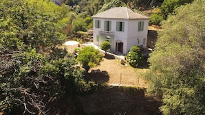 A white building with green shutters, surrounded by lush greenery and hills in the background.