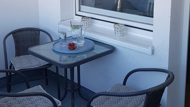 A small balcony with a table and four chairs, a window with a view of buildings, and a glass of water on the table. - Sylter Pirat, Haus Ankerlicht (Sylt)