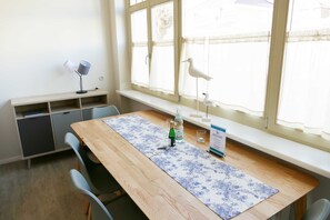 Ein Essbereich mit einem Holztisch, Stühlen, einem Fenster mit Vorhängen, einer kleinen Ablage mit einer Lampe und einem dekorativen Vogel.
