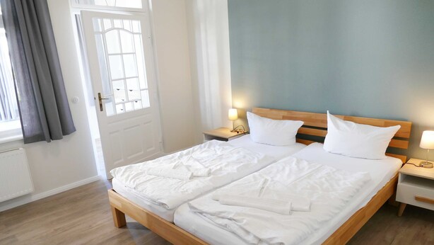 A double bed with white linens, wooden headboard, bedside tables with lamps, and a view of the outdoors through a window with curtains.