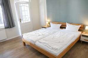 Cama doble con ropa blanca, cabecera de madera, mesitas de noche con lámparas y vista al exterior a través de una ventana con cortinas.