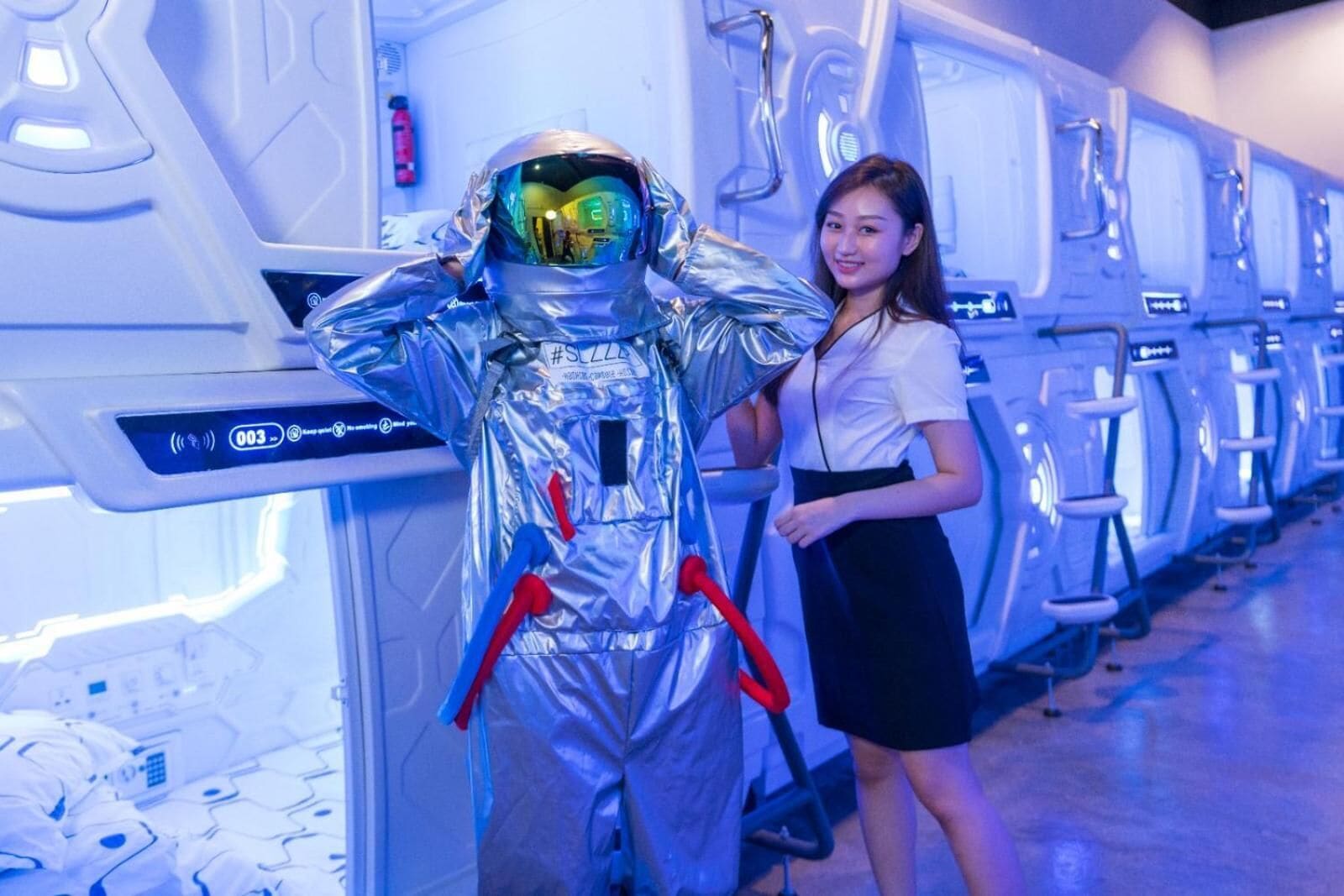 A person in a metallic astronaut suit standing next to a row of white, futuristic-looking pods.