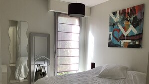 Un dormitorio con cama, armario, espejo, ventana con persianas y un cuadro en la pared.