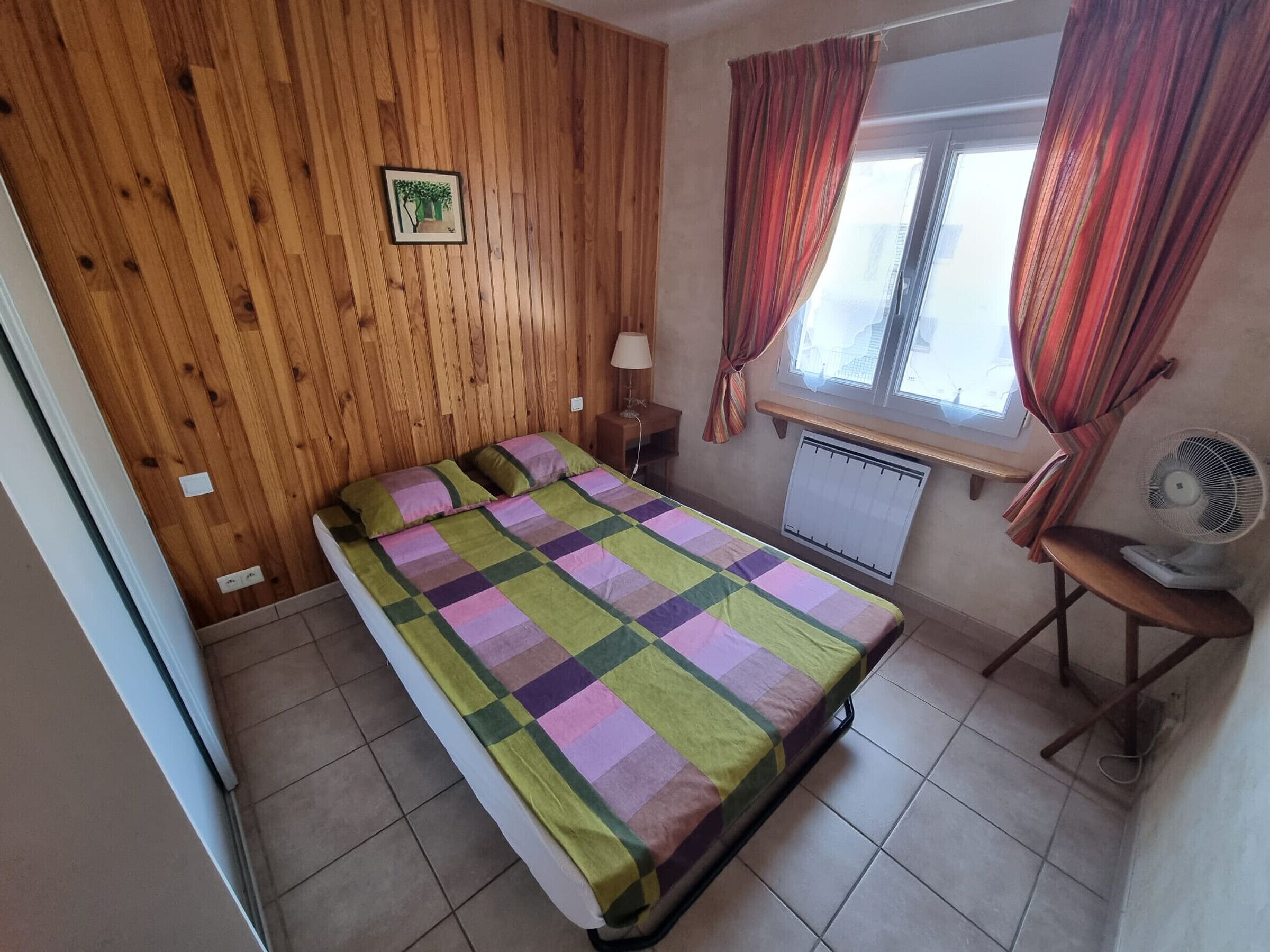 Un dormitorio con cama, ventana con cortinas, radiador, escritorio con lámpara y un cuadro colgado en la pared.