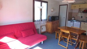 Un espace de vie compact comprenant un canapé rouge, une table à manger en bois avec des chaises, un coin cuisine et une fenêtre avec une lampe blanche.