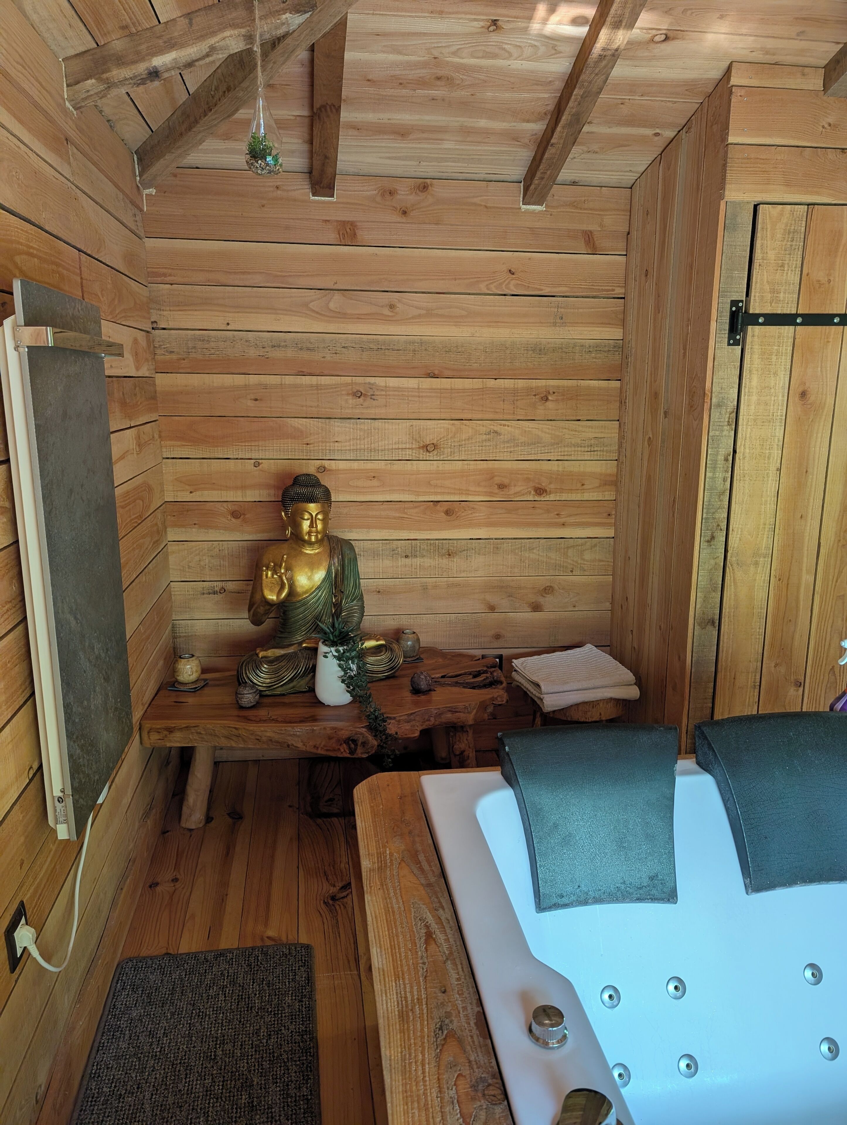 Une pièce de cabane en bois comprenant un lit, une petite table sur laquelle est posée une statue de Bouddha, une lampe suspendue et un plafond en bois.