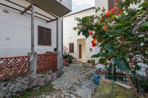 Un étroit sentier pavé de pierres menant à un bâtiment blanc orné de volets bruns, d’une clôture en treillis rouge et d’un arbre aux fleurs rouges.