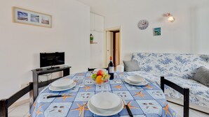 Un coin repas avec une table dressée pour quatre personnes, un téléviseur posé sur un meuble, un canapé à motifs floraux et une horloge fixée au mur.