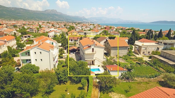 Houses with red-tiled roofs are nestled among lush greenery, set against a mountainous backdrop and the sea.