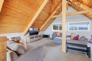 A spacious loft-style living room with a sloped ceiling, wooden beams, a flat-screen TV, and a large window offering a view of the outdoors. - Beach Dreamer-Updated, beautiful beach views (Pacific City)