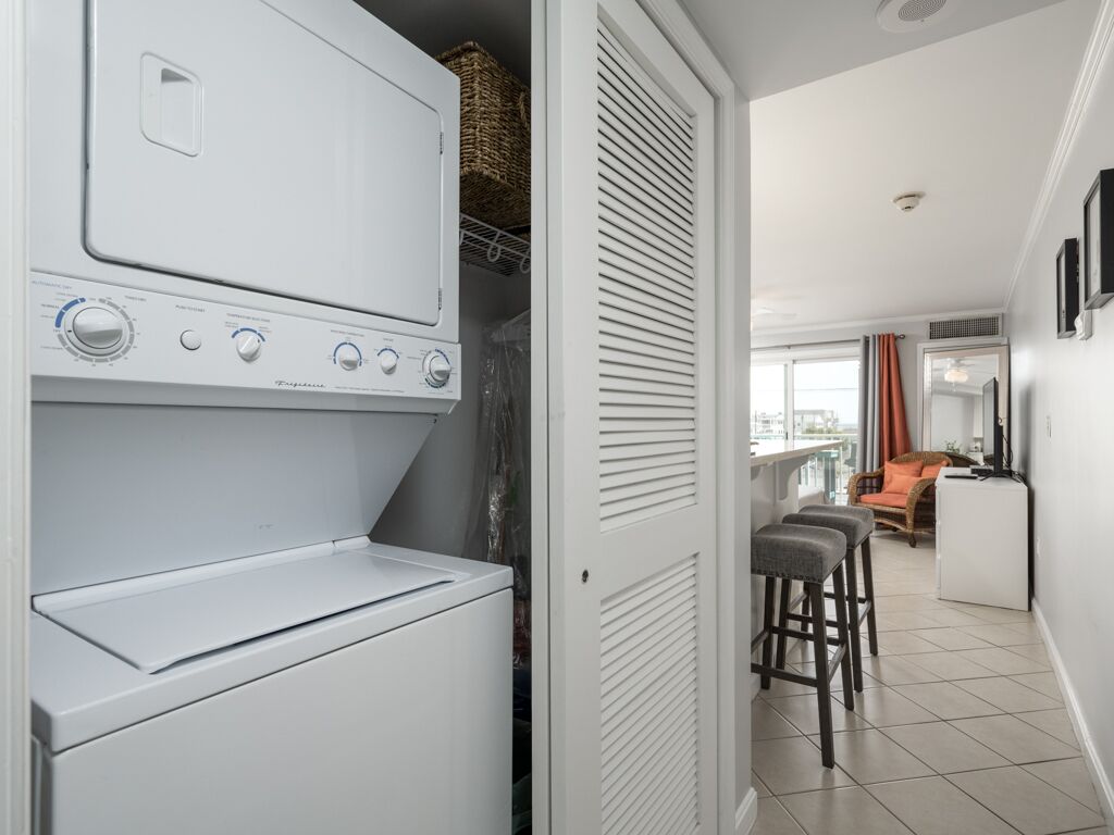 A laundry room with a front-loading washer and dryer, a wicker basket on the dryer, and a sliding door closet. There are two bar stools and a small table in the adjacent living area.