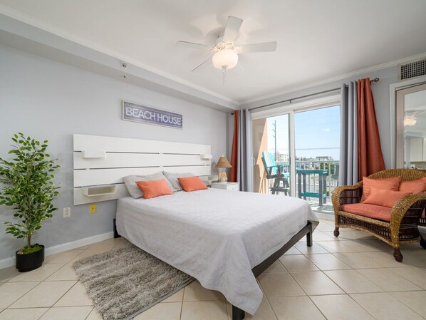 Un dormitorio con cama, ventilador de techo, planta en maceta, silla de mimbre y balcón con vistas.