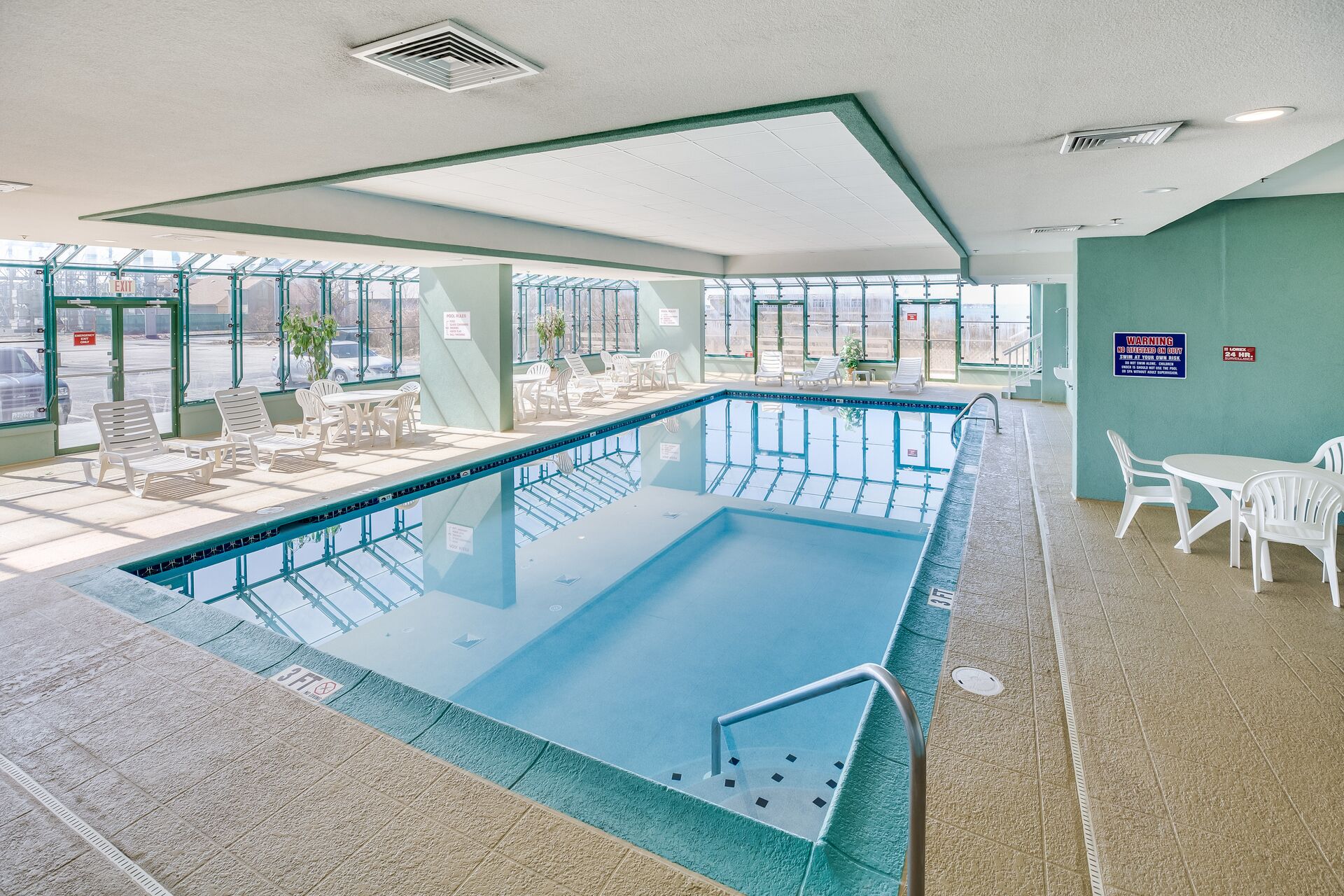 An indoor pool area with clear glass walls, white lounge chairs, and a safety sign on the wall.