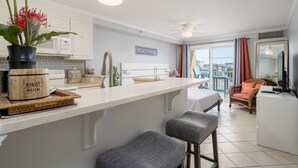 Une cuisine moderne avec un comptoir blanc, deux tabourets de bar, un ventilateur de plafond et un panneau indiquant « MAISON DE PLAGE ».