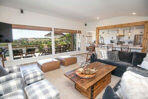 Un vaste espace de vie doté d’une grande fenêtre donnant sur un paysage pittoresque, d’un coin salon confortable et d’une cuisine moderne visible grâce à l’aménagement ouvert.